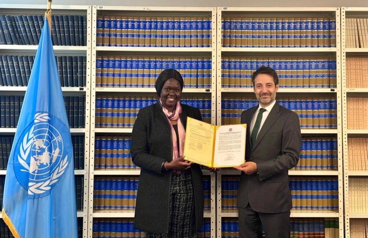 Cecilia Adeng and David Nanopoulos, both smiling proudly, stand in front of a large bookcase holding up a document folder, with a UN flag standing to the side.