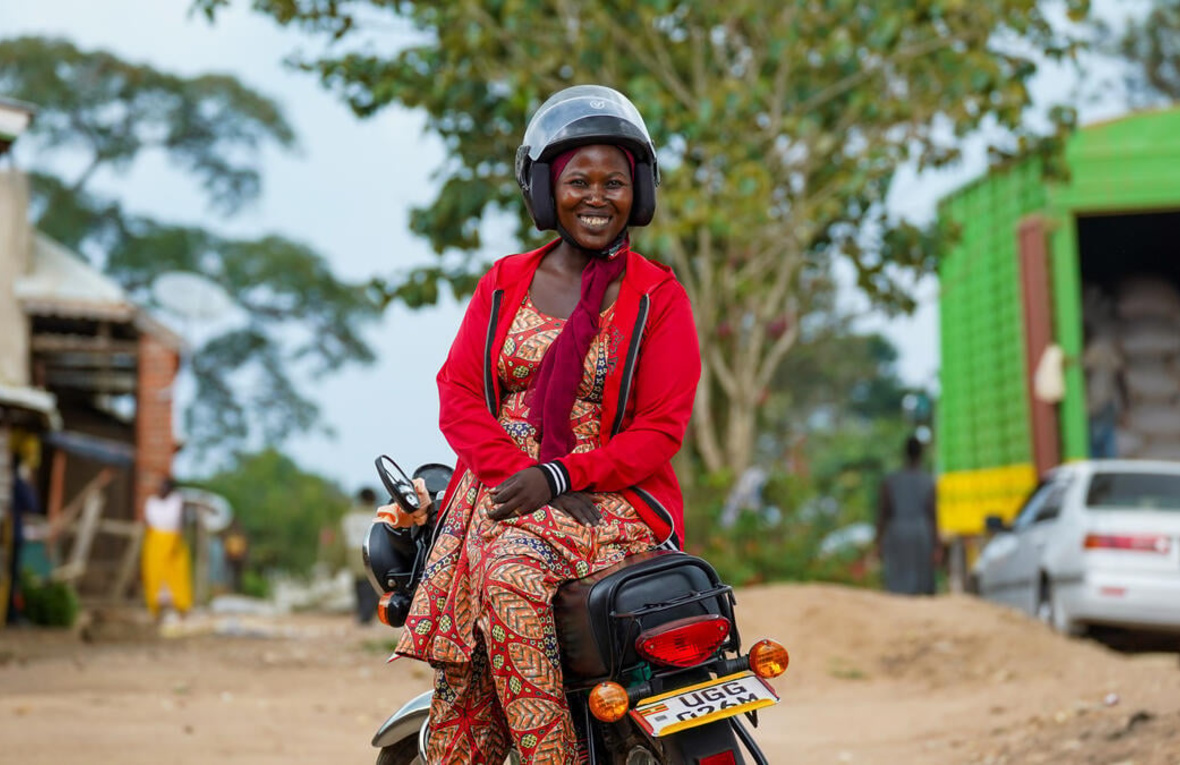 A woman sits sideways on a motorbike saddle wearing a helmet and smiling.