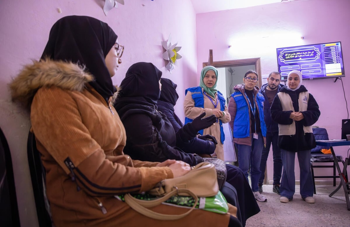 Women sit in a row at a community centre while UNHCR staff stand nearby, speaking to them