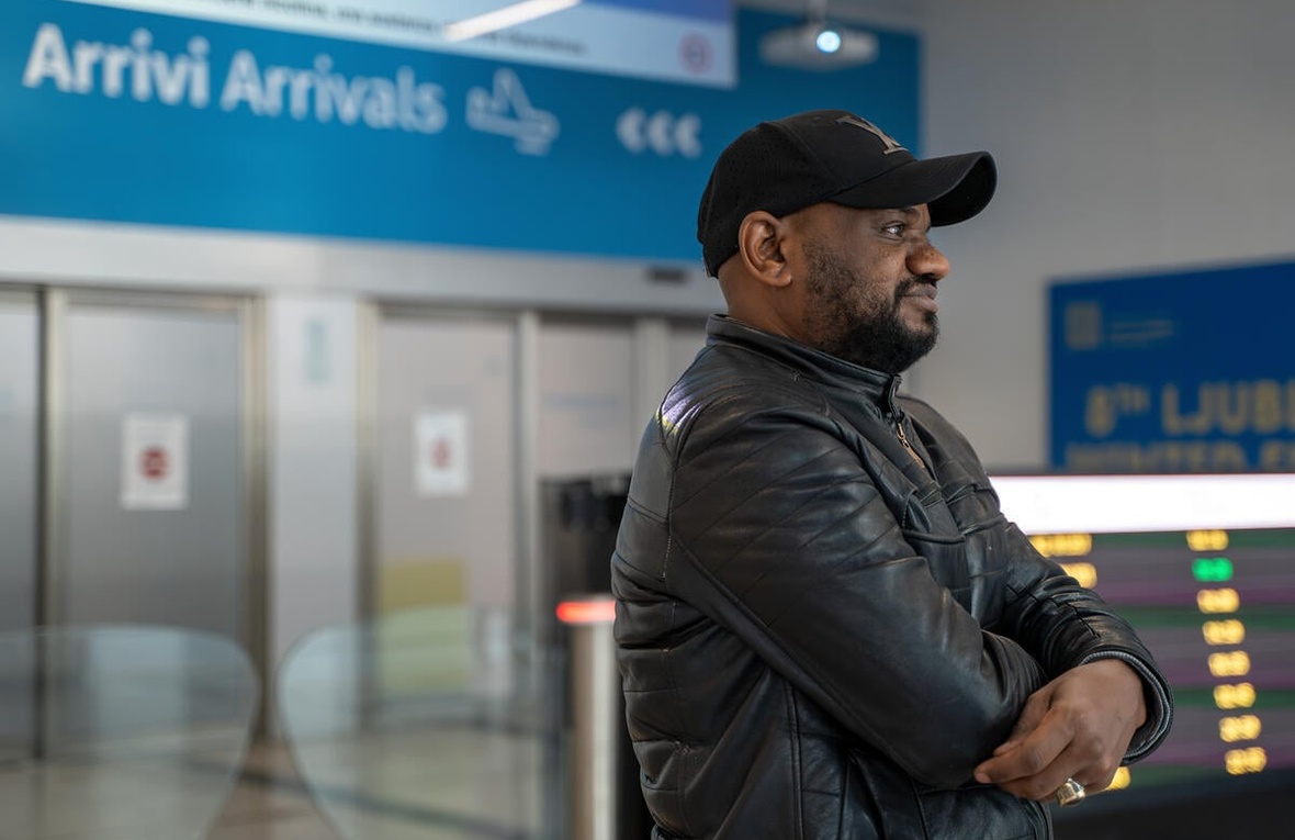 A man wearing a black jacket and cap smiles into the distance in an airport with the Arrivi/Arrivals sign behind him.