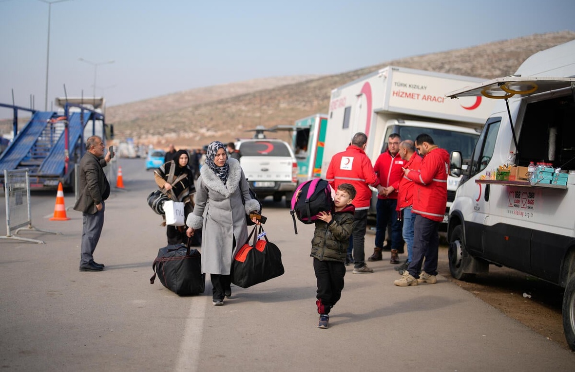 A family carries luggage along a road past vehicles and staff from the 