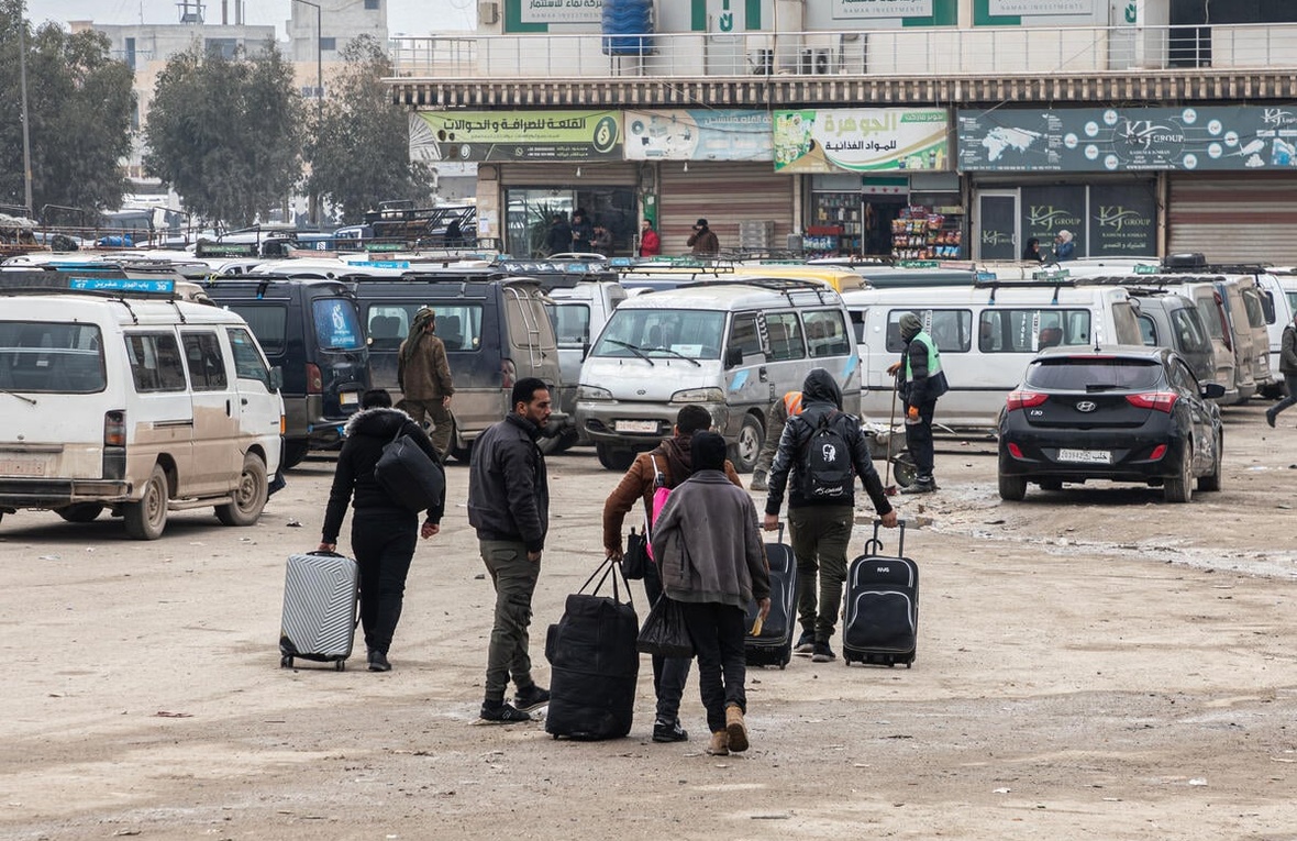 A group of six people walk with suitcases across a dirt parking lot, towards some shops