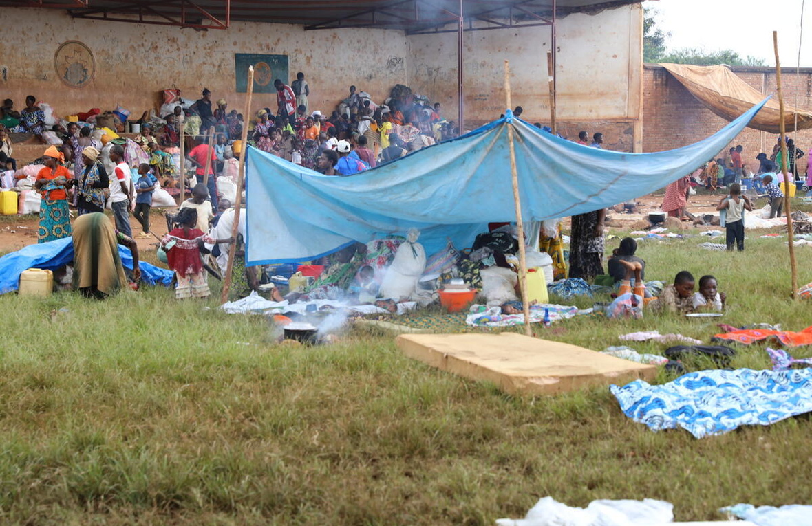 Large groups of people gather with their belongings under a large outdoor structure and smaller makeshift shelters