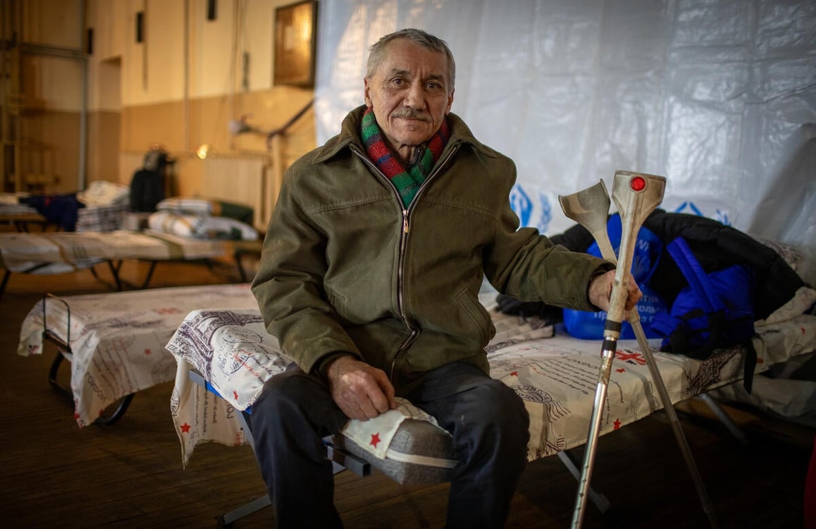 An older man sits on a camp bed in a dormitory, holding some crutches.