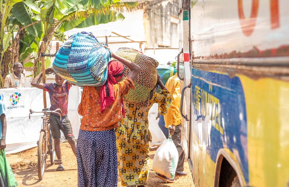 Three Congolese refugees load their belongings onto a bus while a young person stands nearby next to a bicycle, under a banana tree.