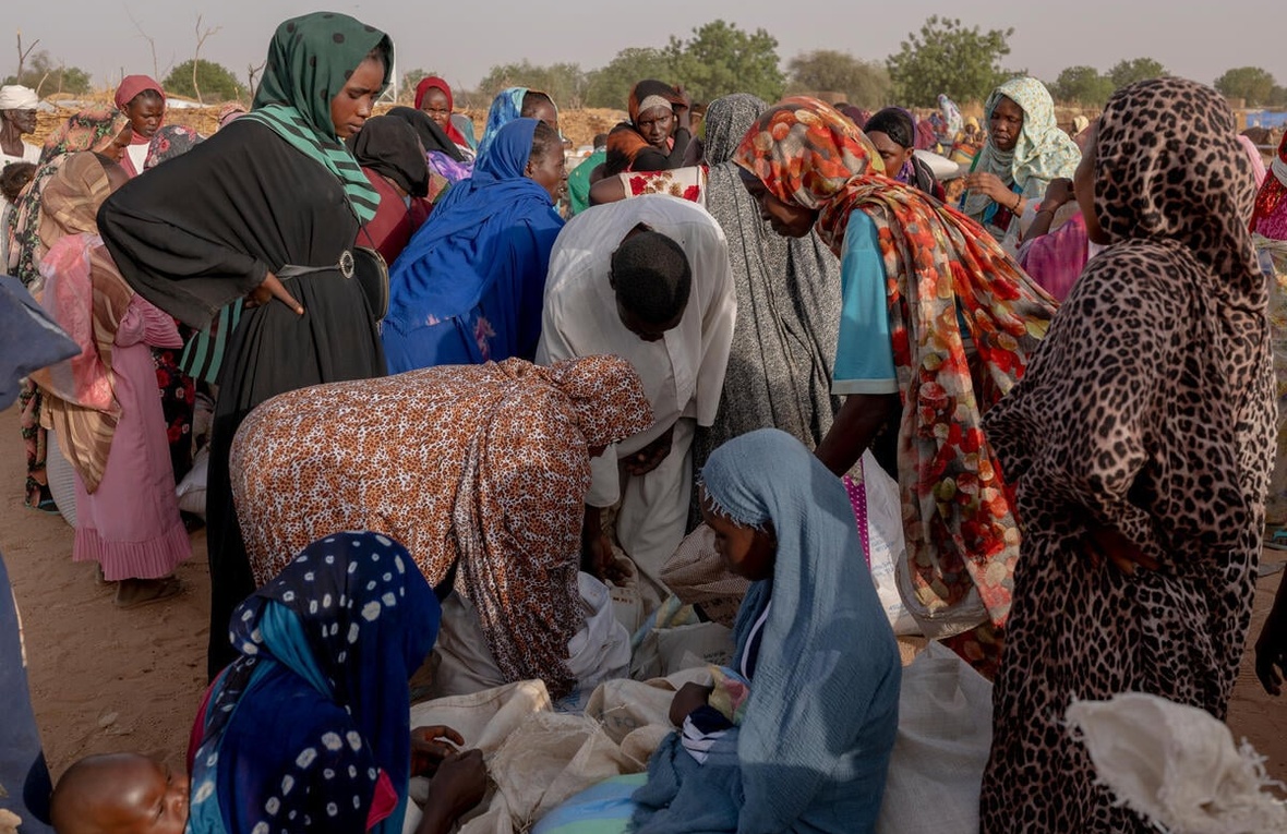 Outside in a sandy expanse with trees in the background, a large group of gather around sacks of grain on the floor