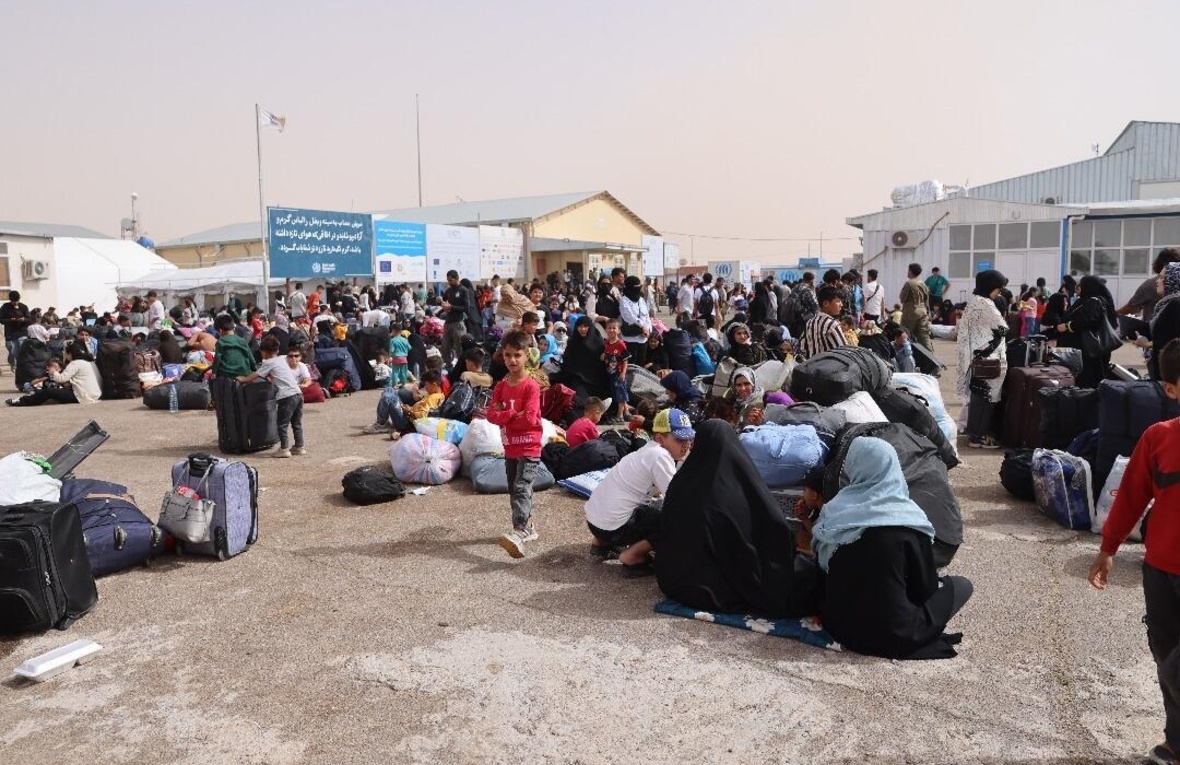 Families with their belongings gather at a border crossing, standing or sitting on the ground.