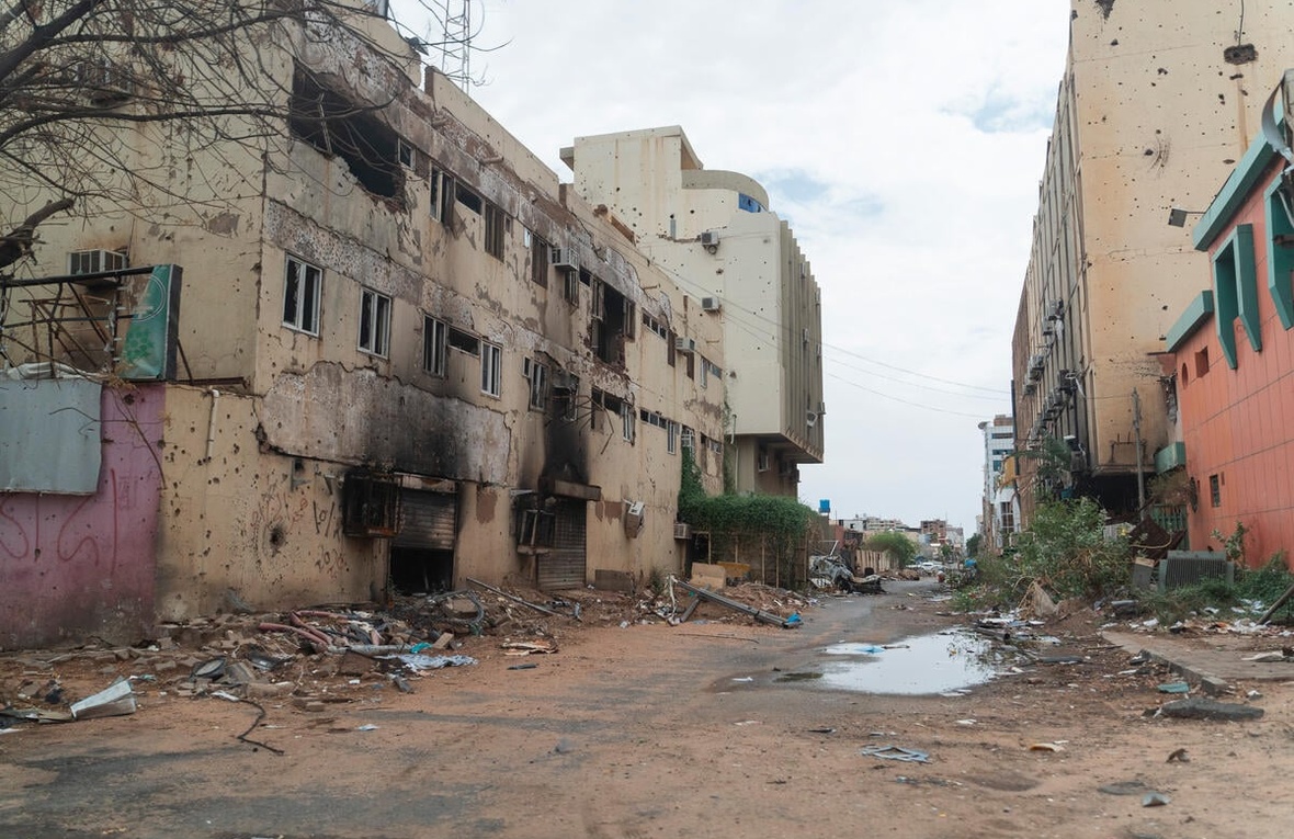 A dirt road with damaged buildings on both sides and building debris along the side of the road
