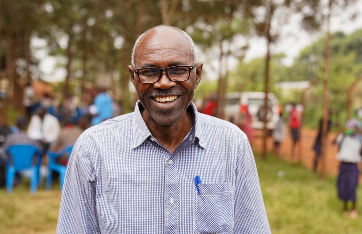 A smiling man wearing a shirt and spectacles stands on a lawn with people and chairs in the background bordered by tall trees