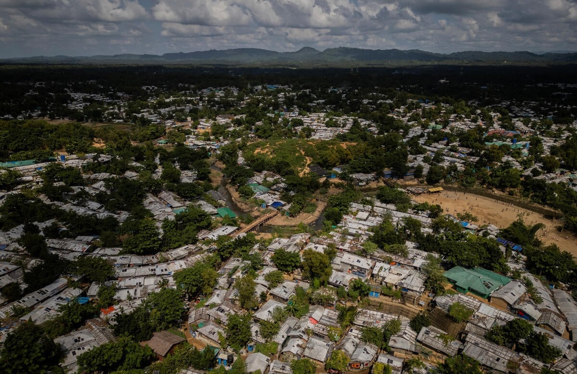 An aerial view of a huge refugee camp, trees throughout, with mountains in the background