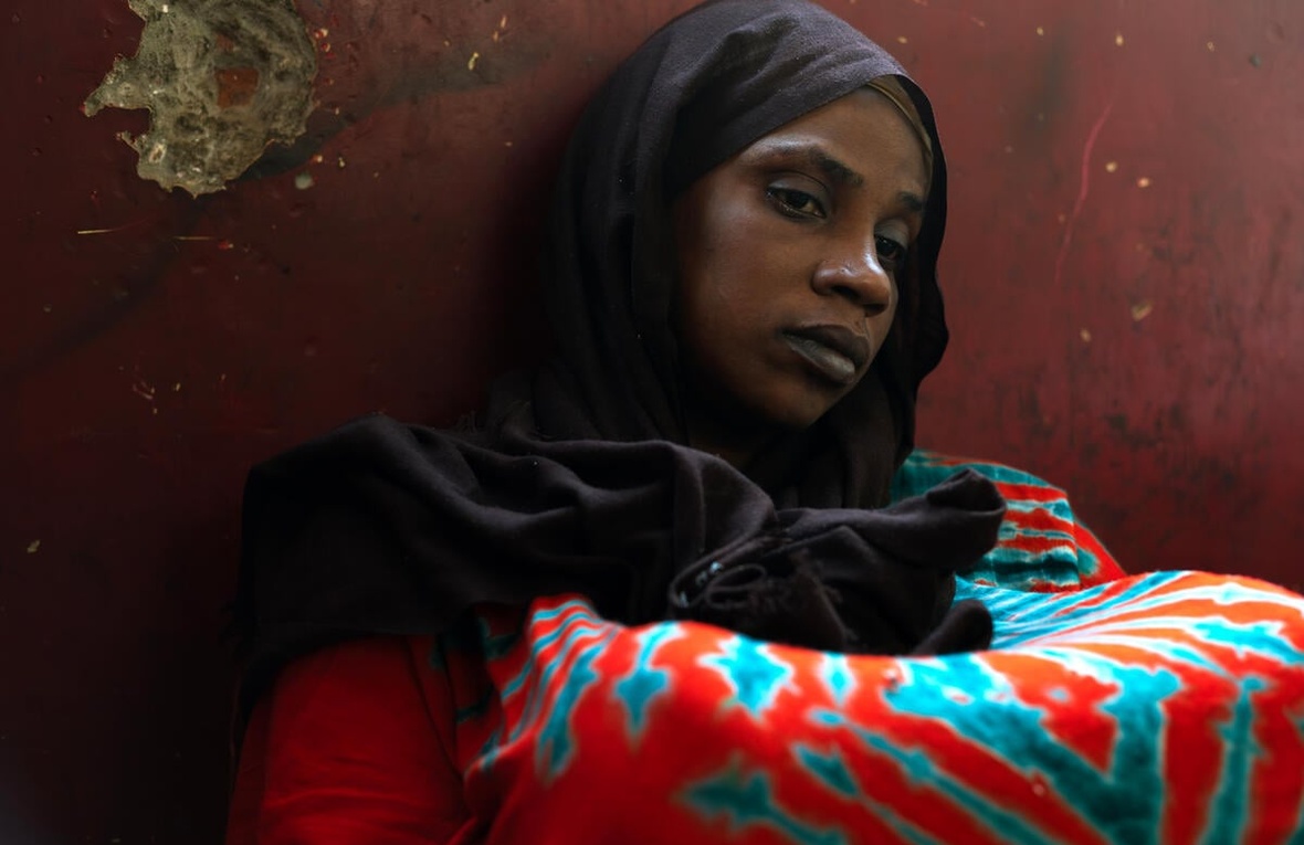 A woman in a colourful dress and a black headscarf sits on the floor leaning against a red-painted, pockmarked wall