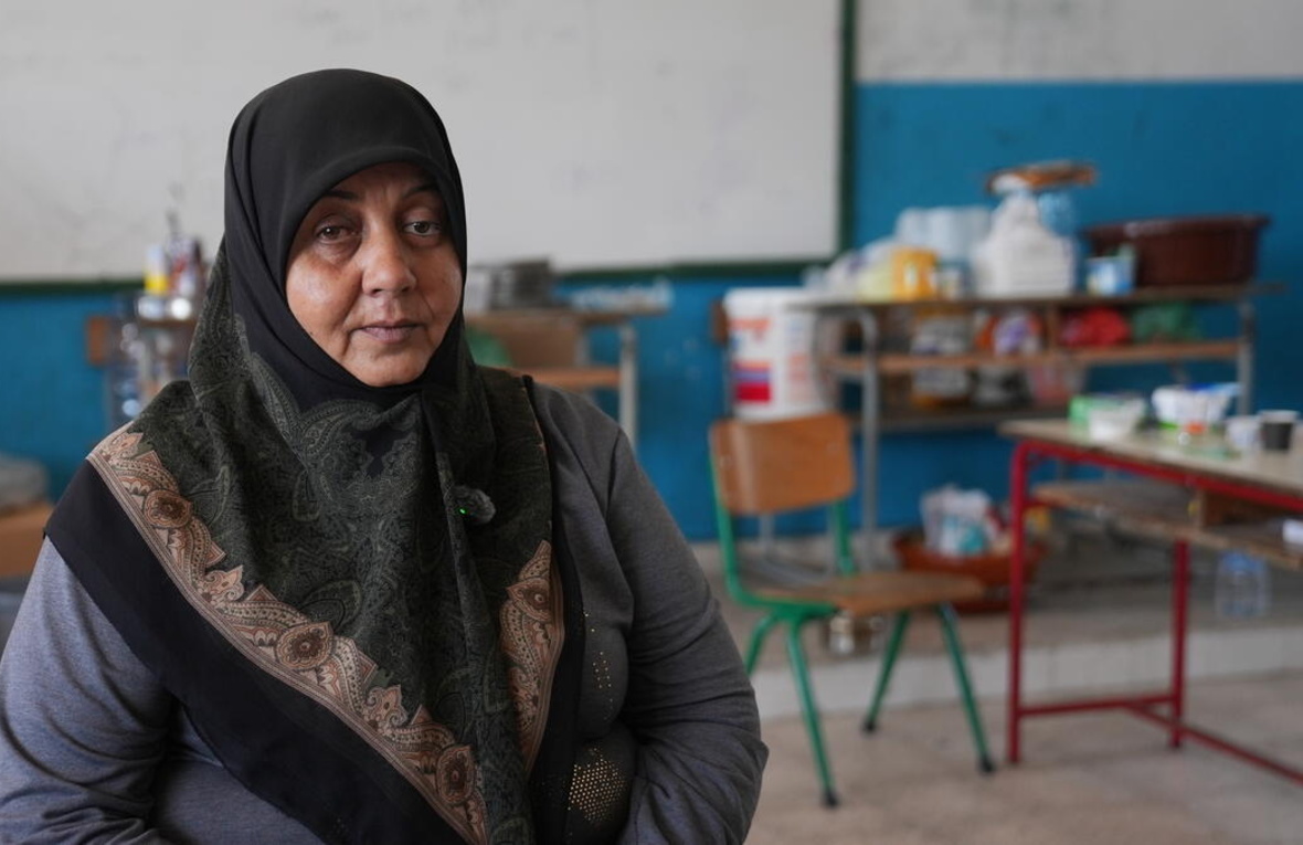 A woman in a headscarf sits in a school classroom with household items on desks in the background