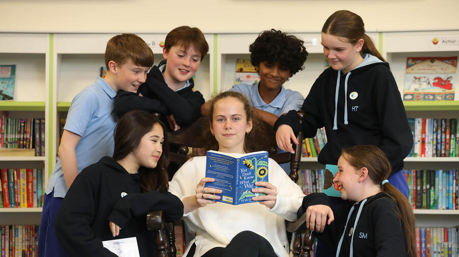 A group of children are gathered around a young woman reading a book who is looking at the camera. 