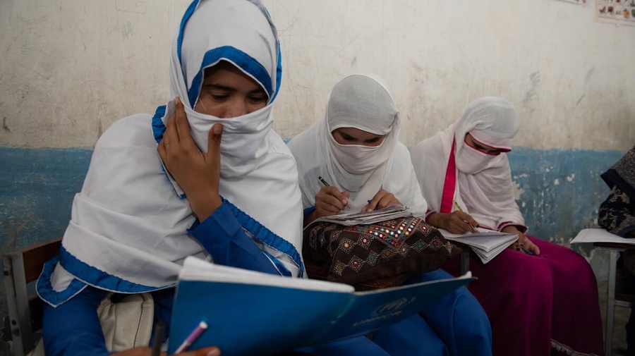 Girls wearing headscarves sit against the wall of a classroom studying their textbooks.
