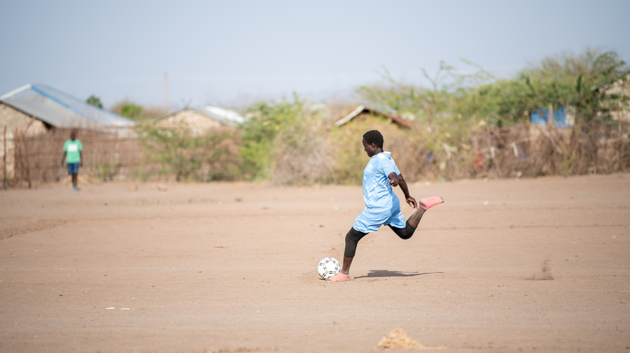 A girl prepares to kick a football on a dusty pitch.