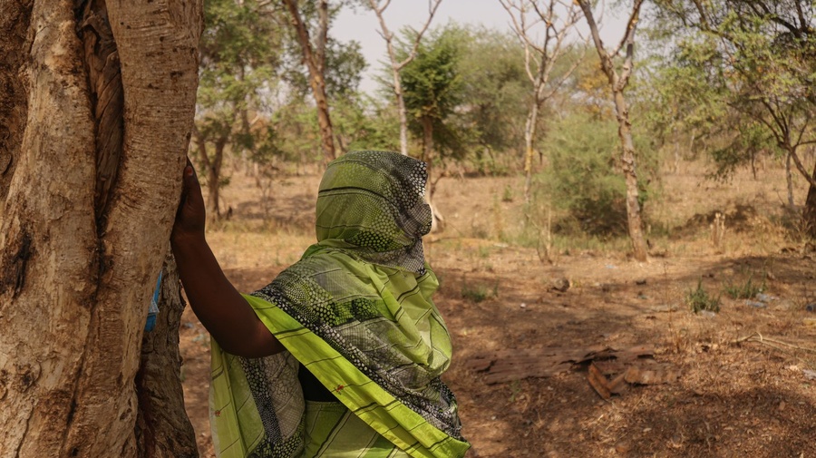 A woman leans against a tree looking out into the scrubland.