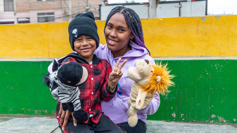 A woman sits outside on brightly-coloured steps with her child on her lap. Both are smiling and holding puppets.