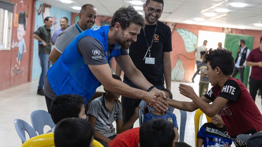 A man wearing a blue vest shakes hands with one of a group of children inside an 