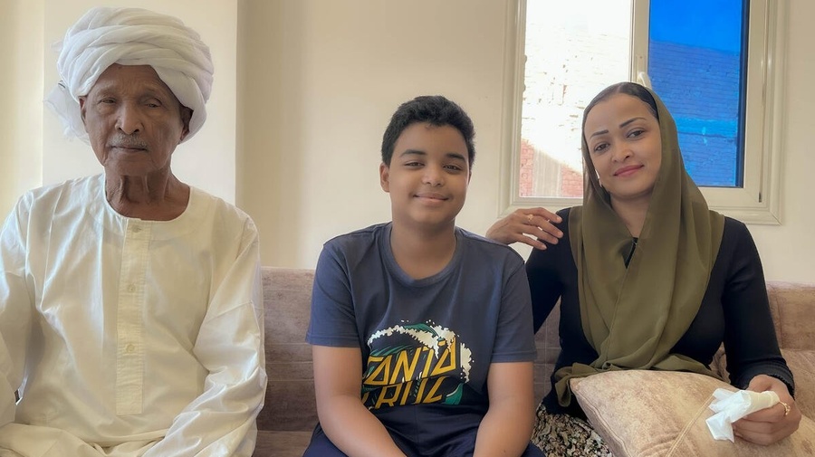 A boy sits on a sofa between his grandfather and mother in their apartment.