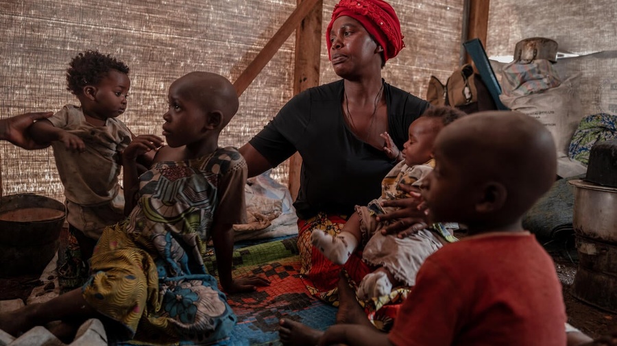A woman sits cradling an infant inside a shelter with three other young children sitting around her.