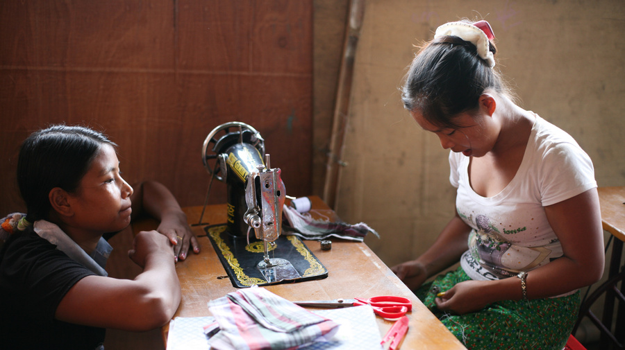 A trainer looks across a sewing machine as her student works hard.
