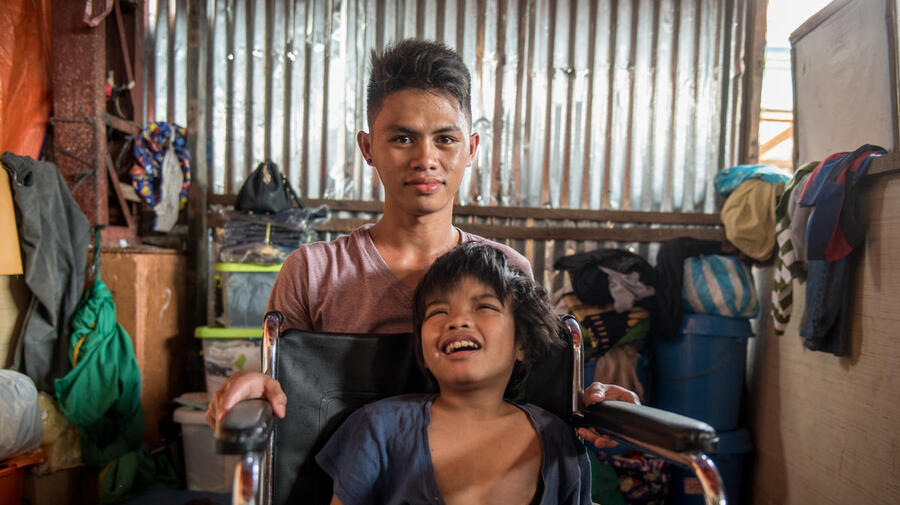 A young man kneels behind a boy who sits in a wheelchair. 