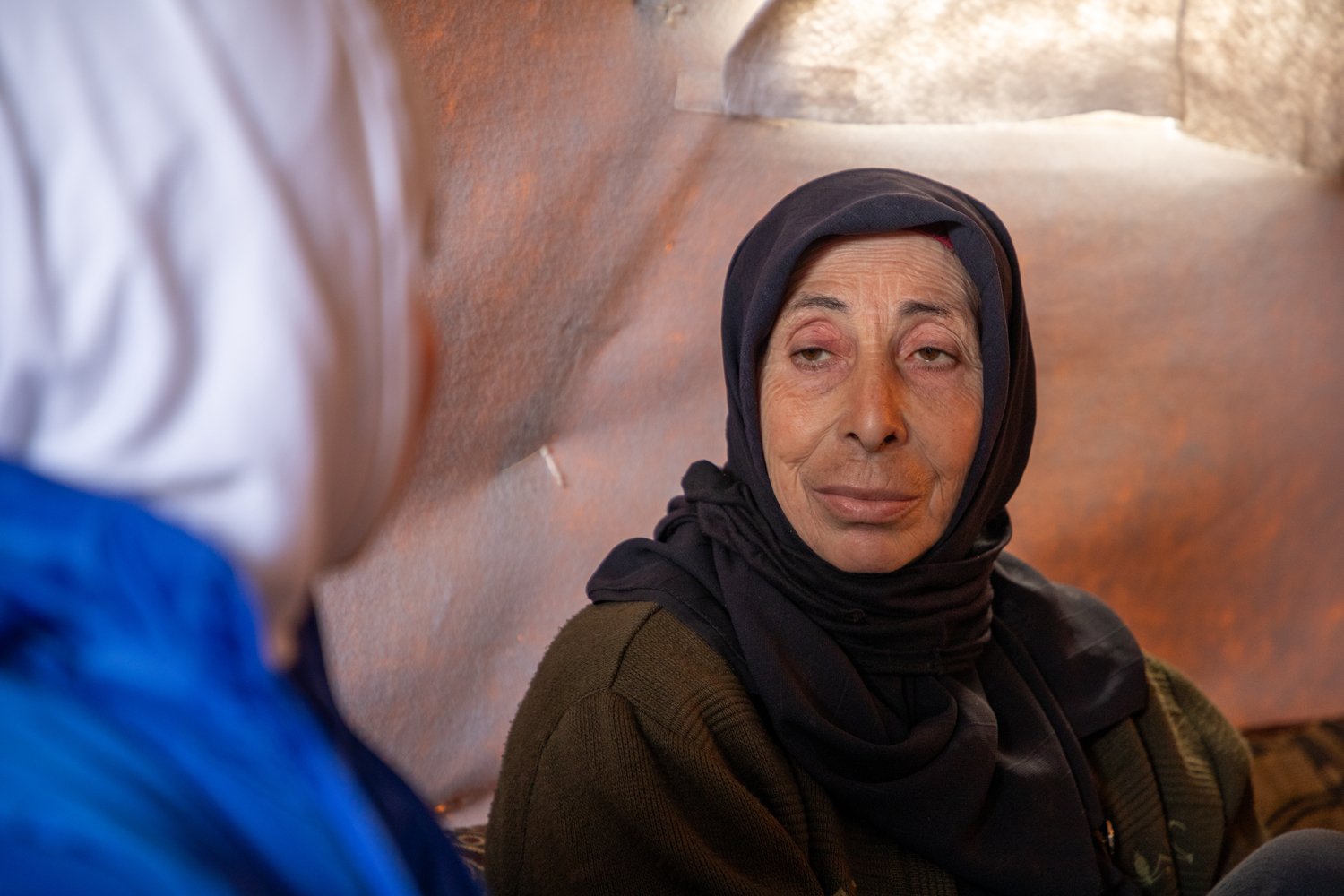 A woman in a dark headscarf sitting inside a tent looks directly at another woman in the foreground wearing a blue jacket and white headscarf whose face is turned away
