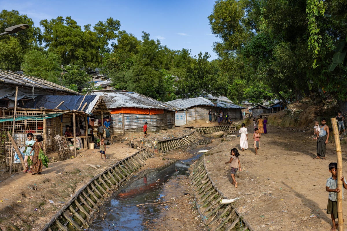 Rohingya refugees walk around a refugee camp beside several shelters and a large trench with some water in it.
