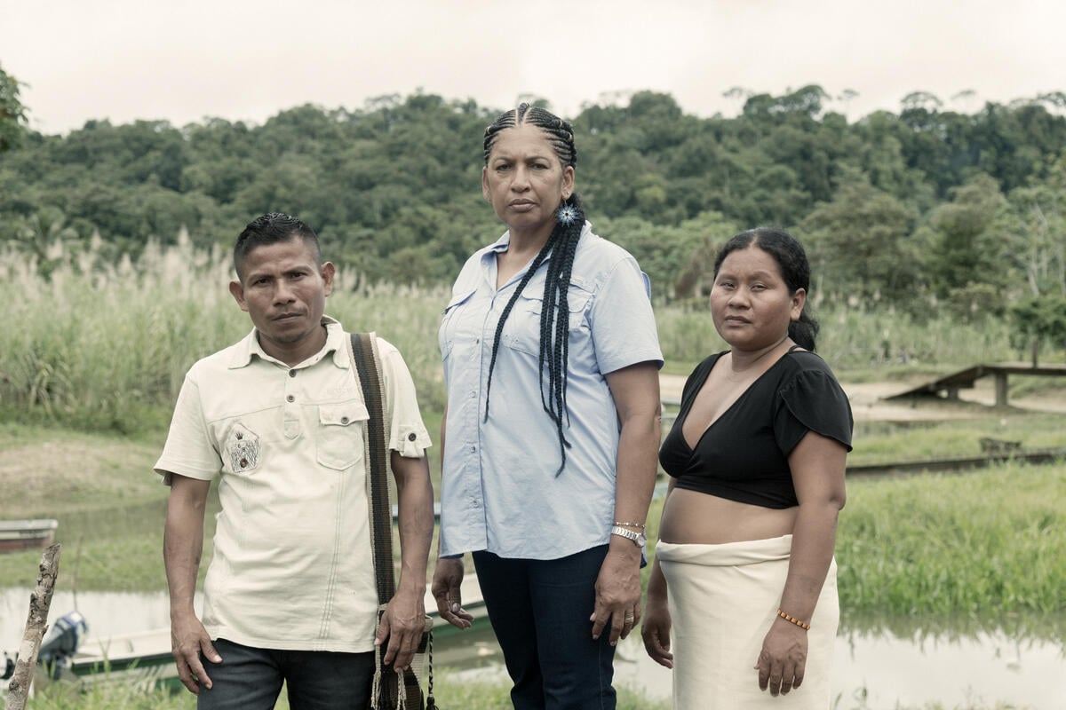 A man and two women stand in front of a stream on the edge of a rainforest.