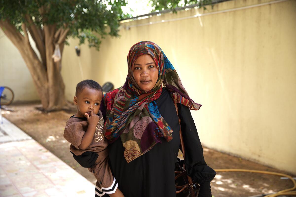 A woman wearing a headscarf stands in a walled courtyard holding a toddler.