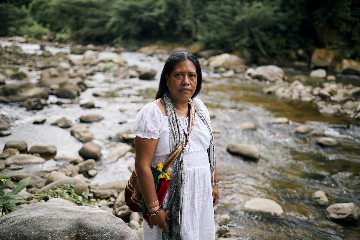A woman wearing a long white dress with a colourful woven bag on her shoulder stands on the bank of a shallow rocky river