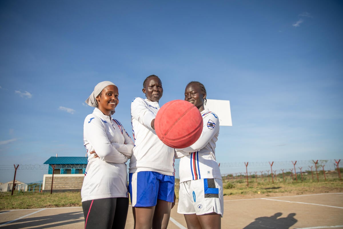 Three women stand together in a basketball court, one holding out a basketball.