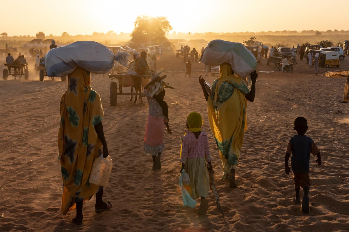 Sudanese refugees and their children walk towards the sun, carrying their belongings on their heads or on carts.