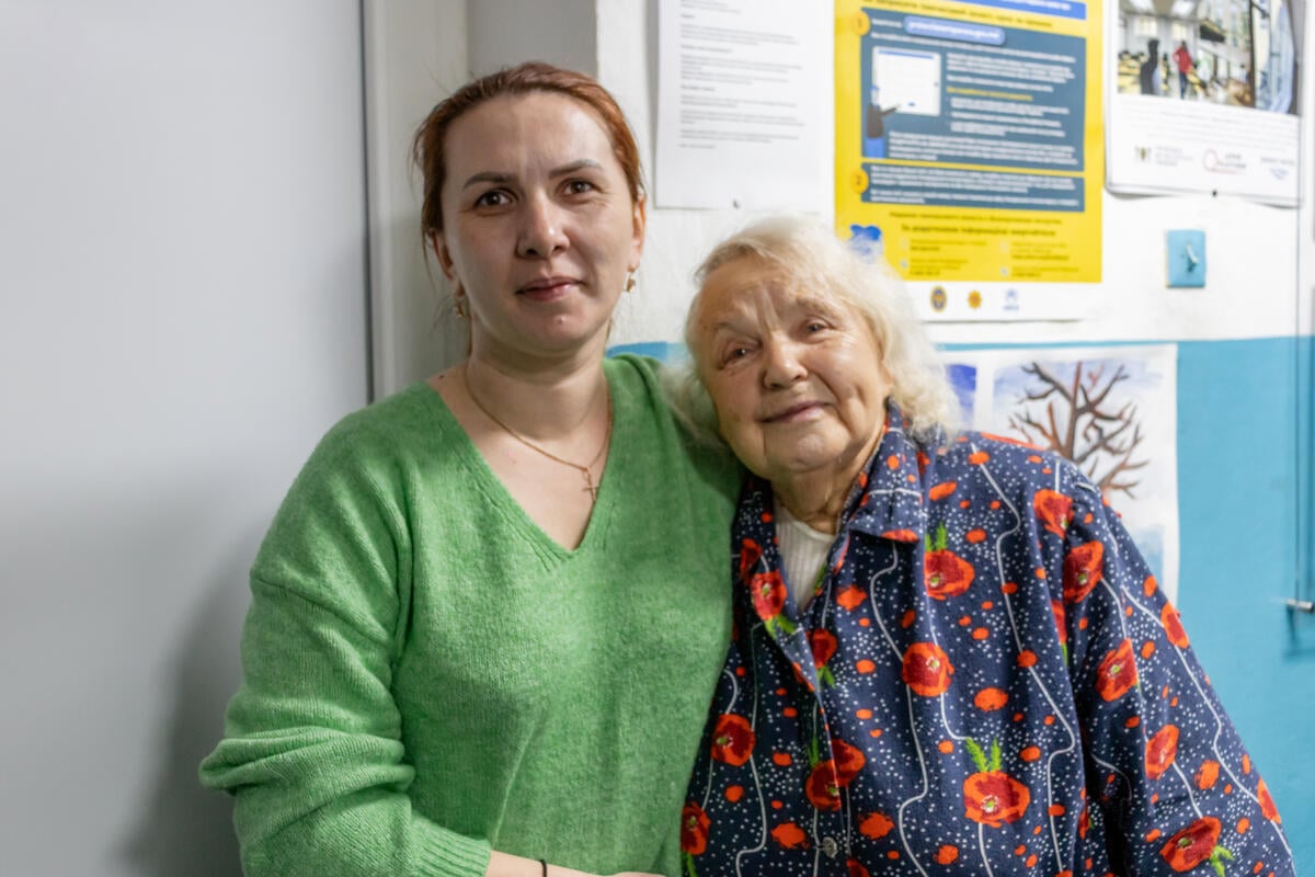 A younger woman and an older woman stand together, arms around each other.