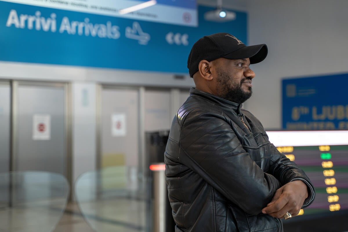 A man wearing a black jacket and cap smiles into the distance in an airport with the Arrivi/Arrivals sign behind him.