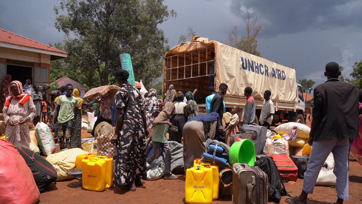 Large groups of people gather with their belongings outside at the back of an open UNHCR truck.