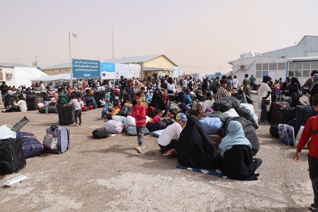 Families with their belongings gather at a border crossing, standing or sitting on the ground.