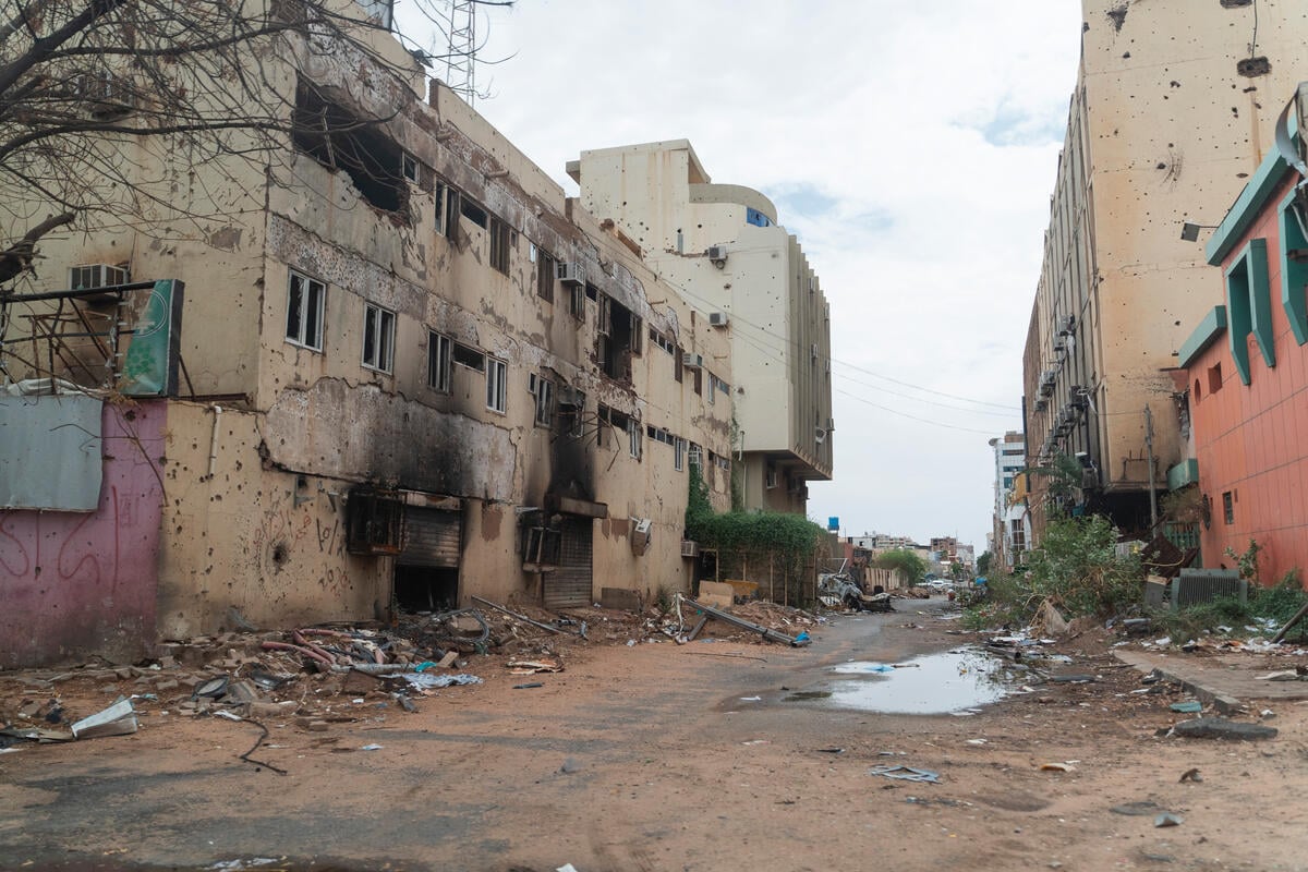 A dirt road with damaged buildings on both sides and building debris along the side of the road