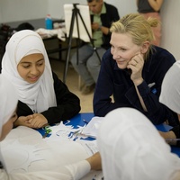 A smiling woman and four girls sit around a table holding scissors and paper 
