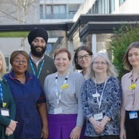 A group of six women and one man, most wearing hospital lanyards and one in a nurse's uniform, stand outside a building flanked by bushes