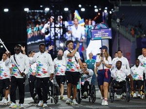 A group of athletes walking and in wheelchairs parade in front of a giant screen display..