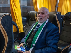 A man sits on a bus wearing a suit and scarf and holding a mobile phone