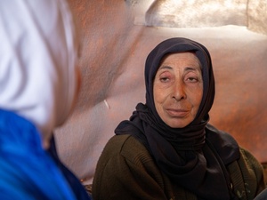 A woman in a dark headscarf sitting inside a tent looks directly at another woman in the foreground wearing a blue jacket and white headscarf whose face is turned away