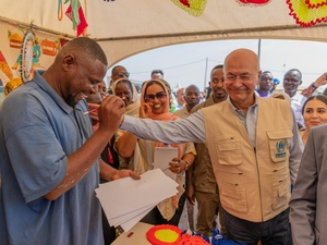 A smiling man drawing a pencil portrait on paper receives a pat on the shoulder from his smiling subject in a tent full of onlookers