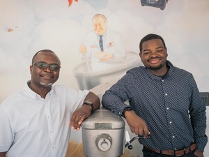 Two men stand either side of an industrial cake mixing machine in front of a mural-painted wall