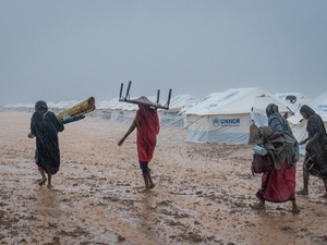 Women carry belongings in pouring rain across muddy ground, with UNHCR tents in the background.