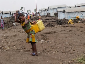 A young girl with two jerry cans on her back stands in front of a row of tents covered in tarpaulin.