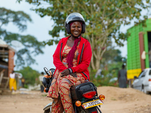 A woman sits sideways on a motorbike saddle wearing a helmet and smiling.