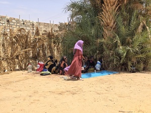 A Sudanese woman walks by a group of people sitting in the shade of some large trees in a dry environment.