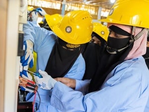 Two women wearing niqabs and yellow hardhats work together to screw a switch to a wall while other women look on.